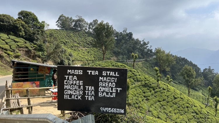 a tea stand with a sign out the front next to rolling green tea plantations in India