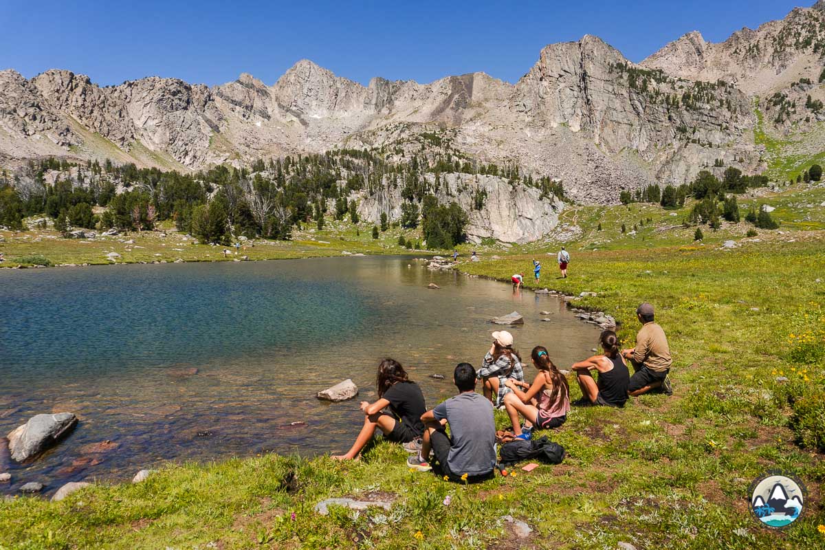 a group of people sitting around the edge of a lake with mountains in the background