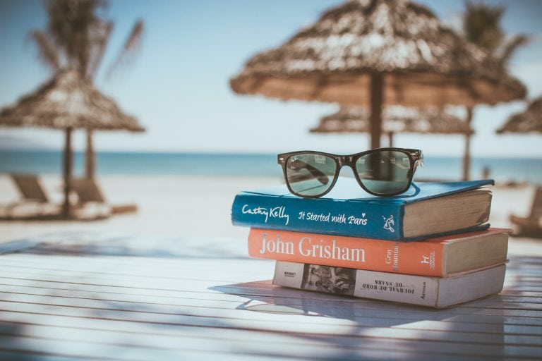 books sitting on a table at the beach with sunglasses on top of the stack and water in the background