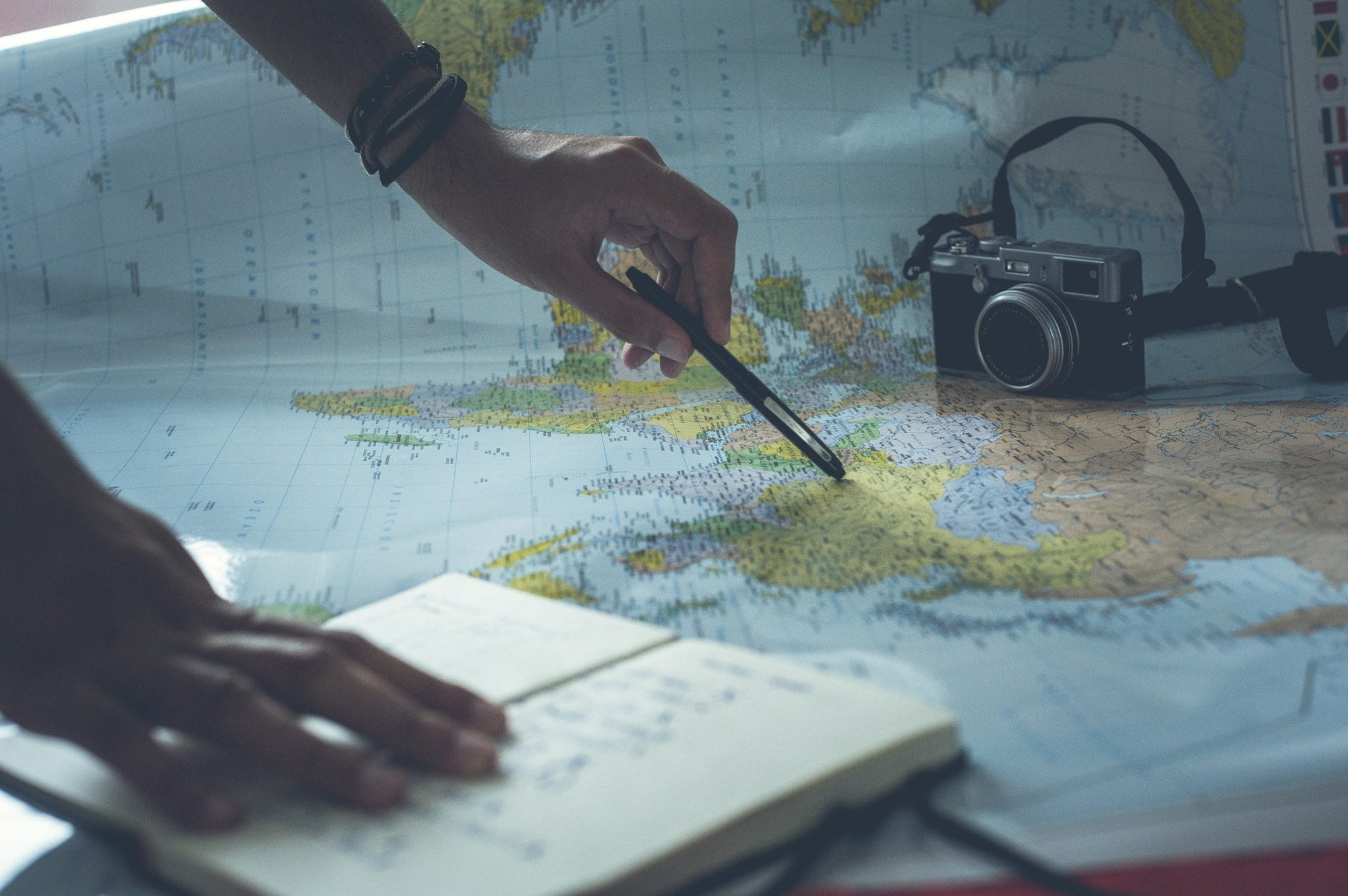 a world map spread out on a table with a camera, journal, and a person pointing to an area with a pen