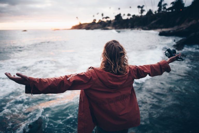 a woman with a red jacket standing looking out to the ocean with her arms spread wide