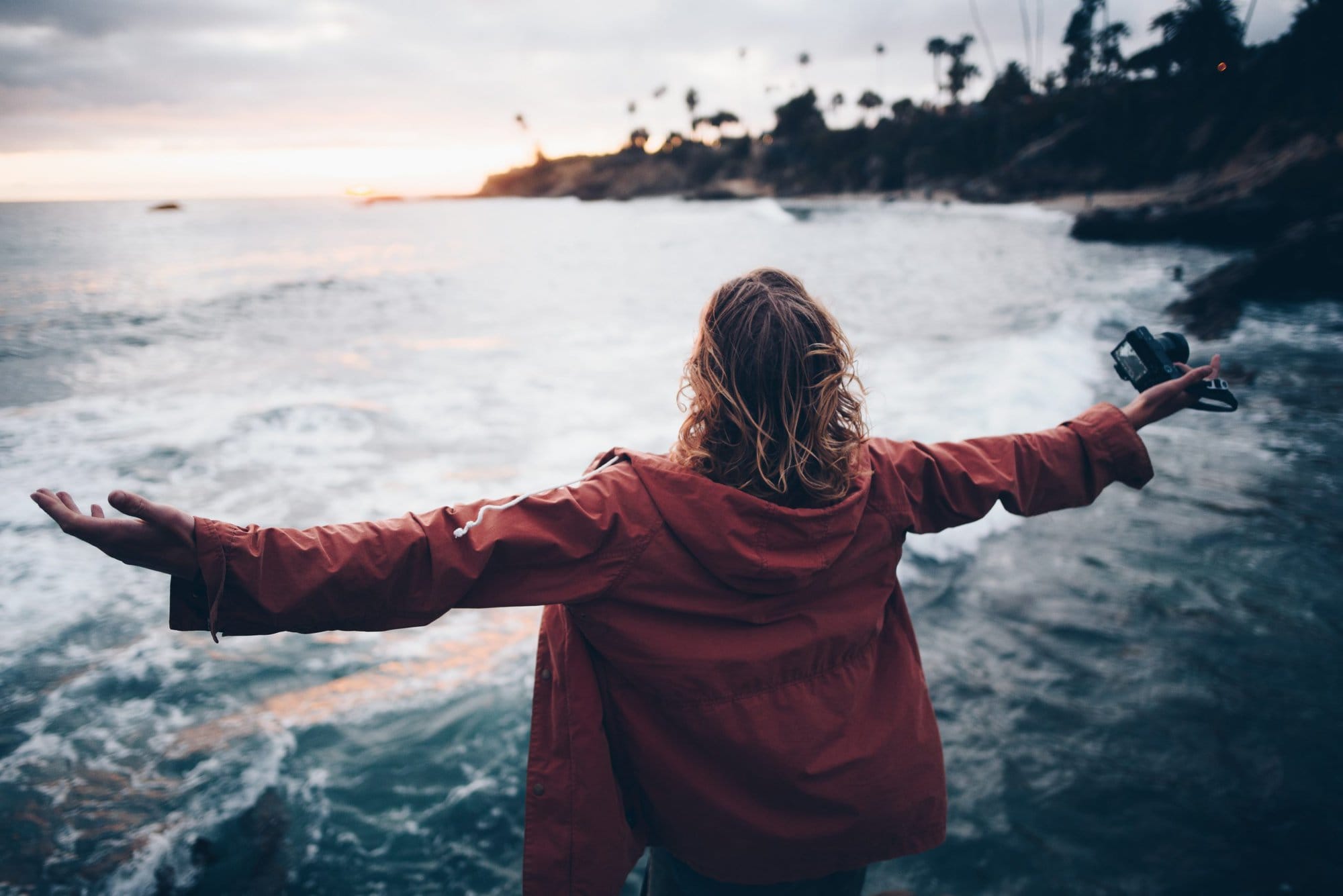 a woman with a red jacket standing looking out to the ocean with her arms spread wide