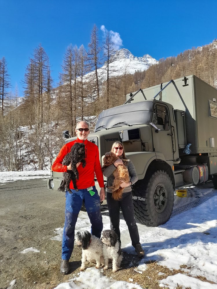 a man, woman and 4 dogs standing in front of a truck in the snow